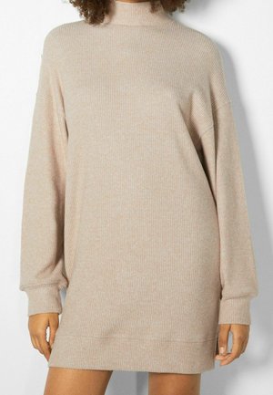 Persona que lleva un vestido de punto de canalé color beige con cuello alto y mangas largas, de pie frente a un fondo claro.