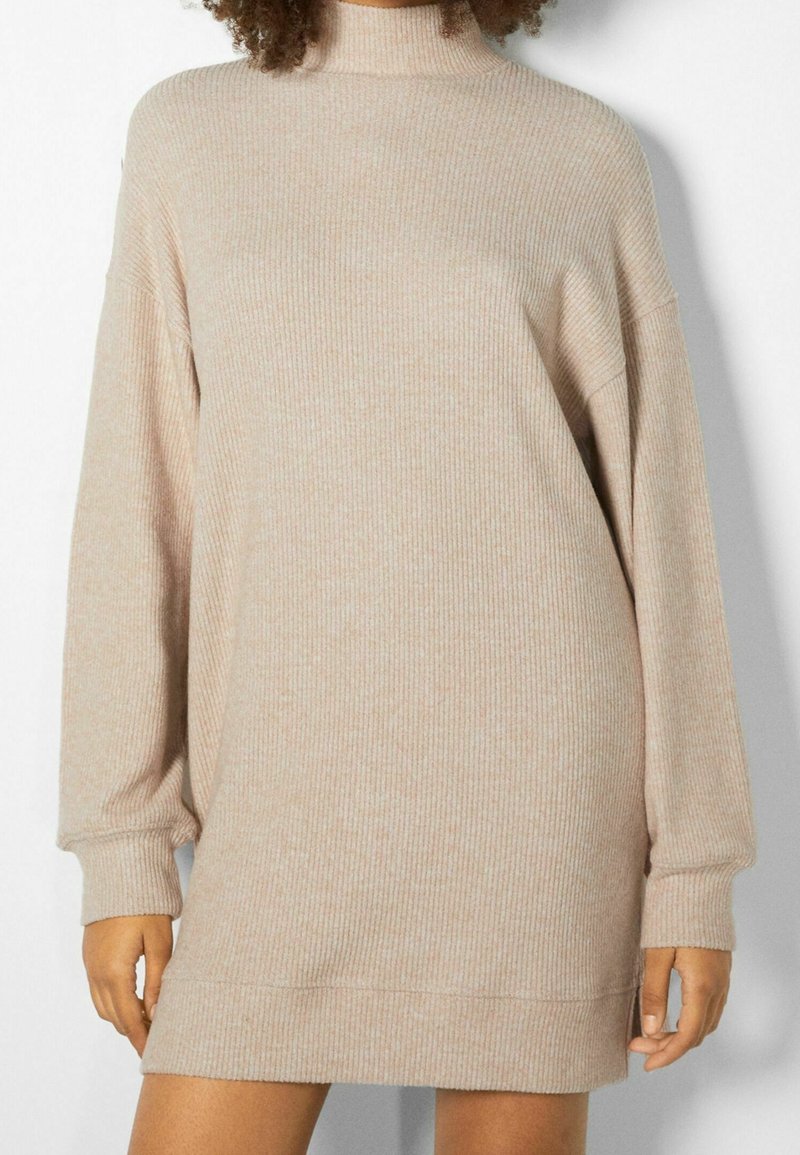 Personne portant une robe-pull à col roulé beige en maille côtelée avec manches longues, debout devant un fond clair.