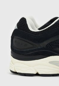 Zapatilla deportiva negra con parte superior de malla y gamuza, mediopié acolchado blanco y panel trasero texturizado con detalle de marca.