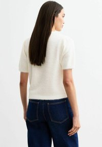 Un pull blanc et duveteux à manches courtes avec un col rond, rentré dans un jean taille haute bleu foncé. Texture douce et détail de bord subtil.