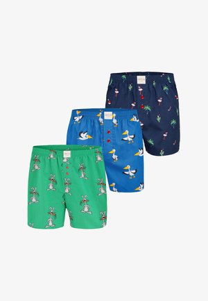 Trois paires de shorts de bain : verts avec des lapins de dessin animé, bleus avec des pélicans et des décorations rouges, et navy avec des flamants roses et des palmiers.