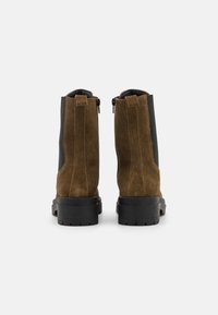 Botas de tornozelo em camurça castanha com um painel lateral elástico preto, fecho de correr traseiro e sola de borracha preta, apresentando detalhes texturizados.