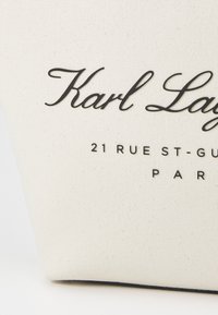 Krämfärgad kanvasväska med svart text som står "Karl Lagerfeld" och adress i Paris, med en smooth yta och robust konstruktion.