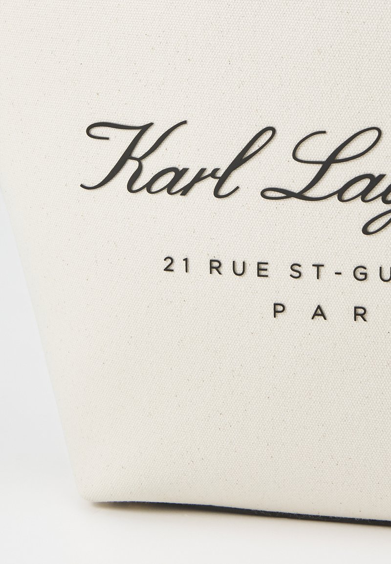 Sac en toile de couleur crème avec du texte noir indiquant "Karl Lagerfeld" et l'adresse de Paris, arborant une texture lisse et une construction robuste.