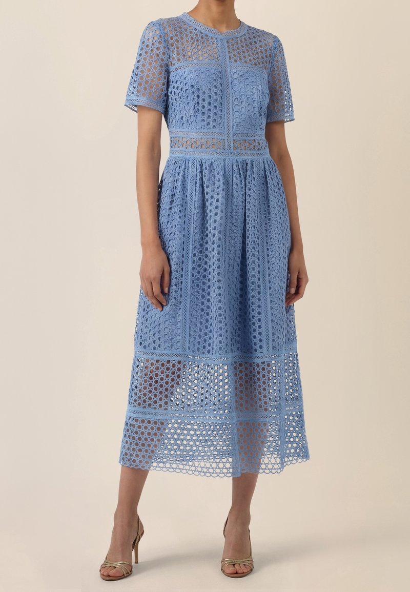 Femme portant une robe en dentelle bleu clair jusqu’aux genoux, à manches courtes, avec des découpes circulaires et des sandales à talons hauts couleur nude.