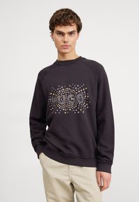 Sudadera gris oscuro con cuello redondo, con un diseño decorativo de tachuelas en oro y plata. Puños de canalé con detalle de dobladillo sin rematar.