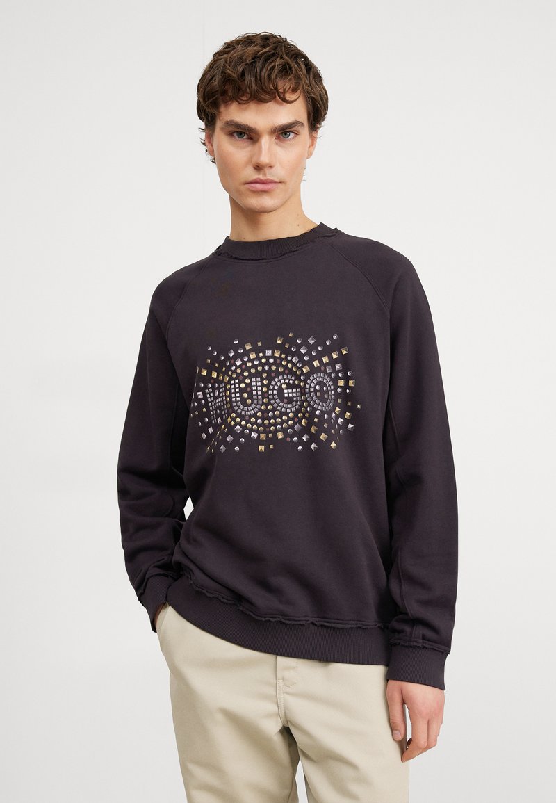 Sudadera gris oscuro con cuello redondo, con un diseño decorativo de tachuelas en oro y plata. Puños de canalé con detalle de dobladillo sin rematar.