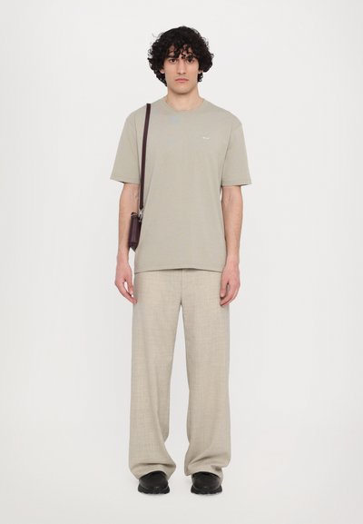 Beige katoenen t-shirt met korte mouwen, gecombineerd met lichtbeige linnen broek en zwarte schoenen. Model houdt een donkerbruin schoudertasje vast.