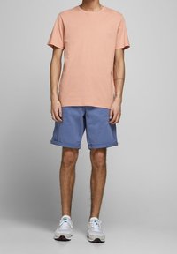 Světle růžové bavlněné tričko s kulatým výstřihem, v kombinaci s modrými kraťasy po kolena a bílými teniskami. Jednoduchý design, bez vzorů.