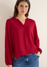 Rød langærmet bluse med V-udskæring og knapdetaljer. Fremstillet af blødt stof med elastiske manchetter. Kombineret med denimshorts.