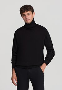 Maglione a collo alto nero in maglia lavorata, con colletto e polsini a coste, indossato da un modello su uno sfondo grigio chiaro.
