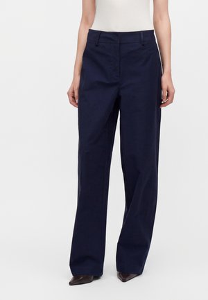 Personne portant un pantalon taille haute à jambes larges bleu marine foncé avec un bouton et une fermeture éclair à l'avant, associé à un haut clair et des chaussures pointues marron.