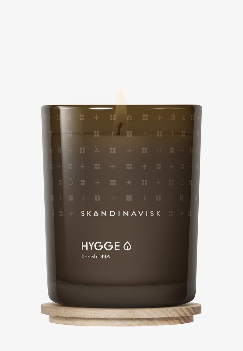 Braunfarbige Glaskerze mit brennendem Docht, verziert mit gravierten Mustern und einem Holzsockel. Das Etikett trägt die Aufschrift "SKANDINAVISK HYGGE Dänisches DNA."