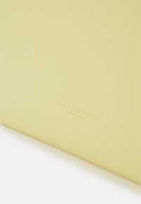 Coin d'un sac Marc O'Polo jaune pâle avec un logo de marque subtilement embossé et un texte sur une surface en cuir lisse.