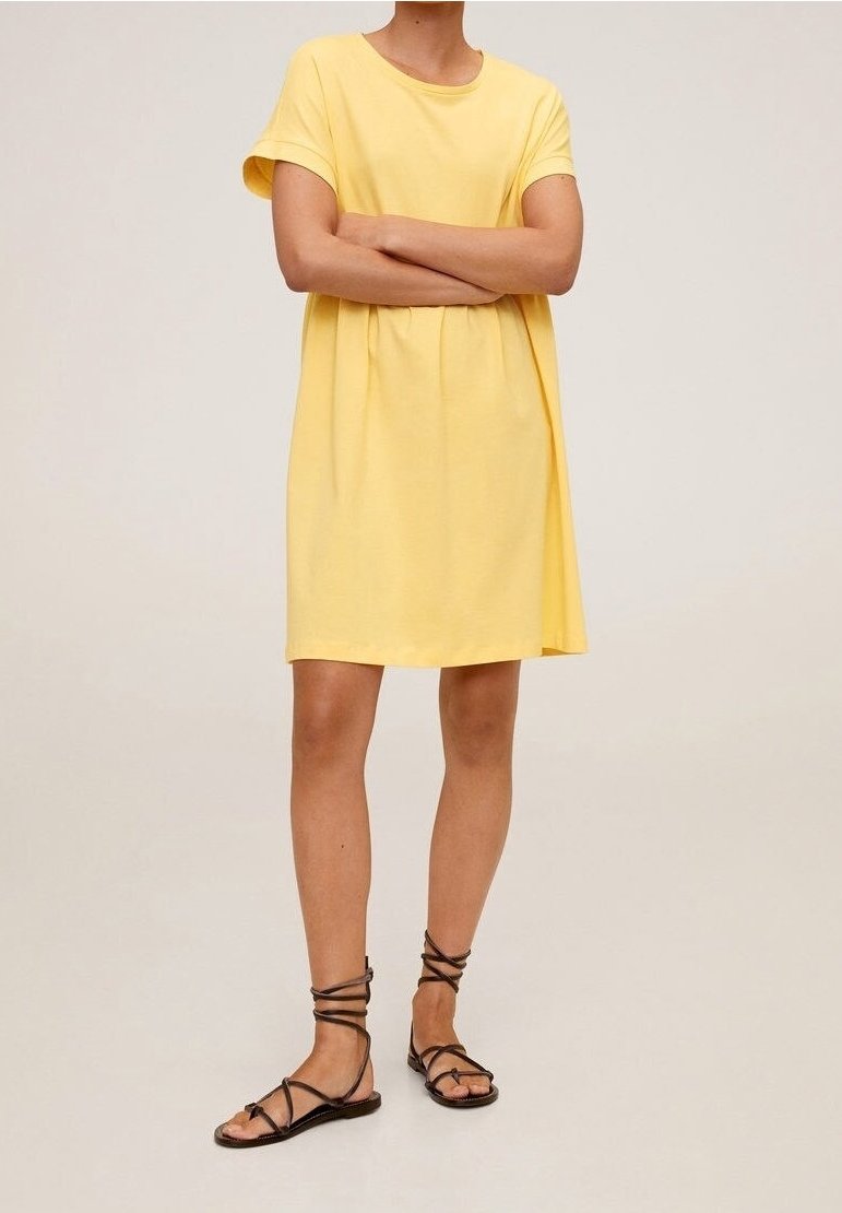 Vestido amarillo de algodón con mangas cortas y un ajuste relajado. Combinado con sandalias negras de tiras, mostrando un diseño sencillo y casual.