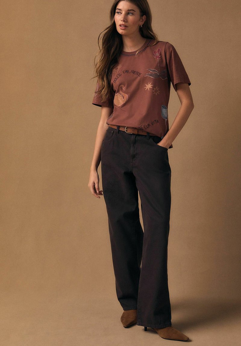 Kobieta ubrana w brązowy T-shirt z grafiką i napisami, czarne spodnie o szerokich nogawkach, brązowy pasek i botki z ostrym czubkiem, stojąca na beżowym tle.