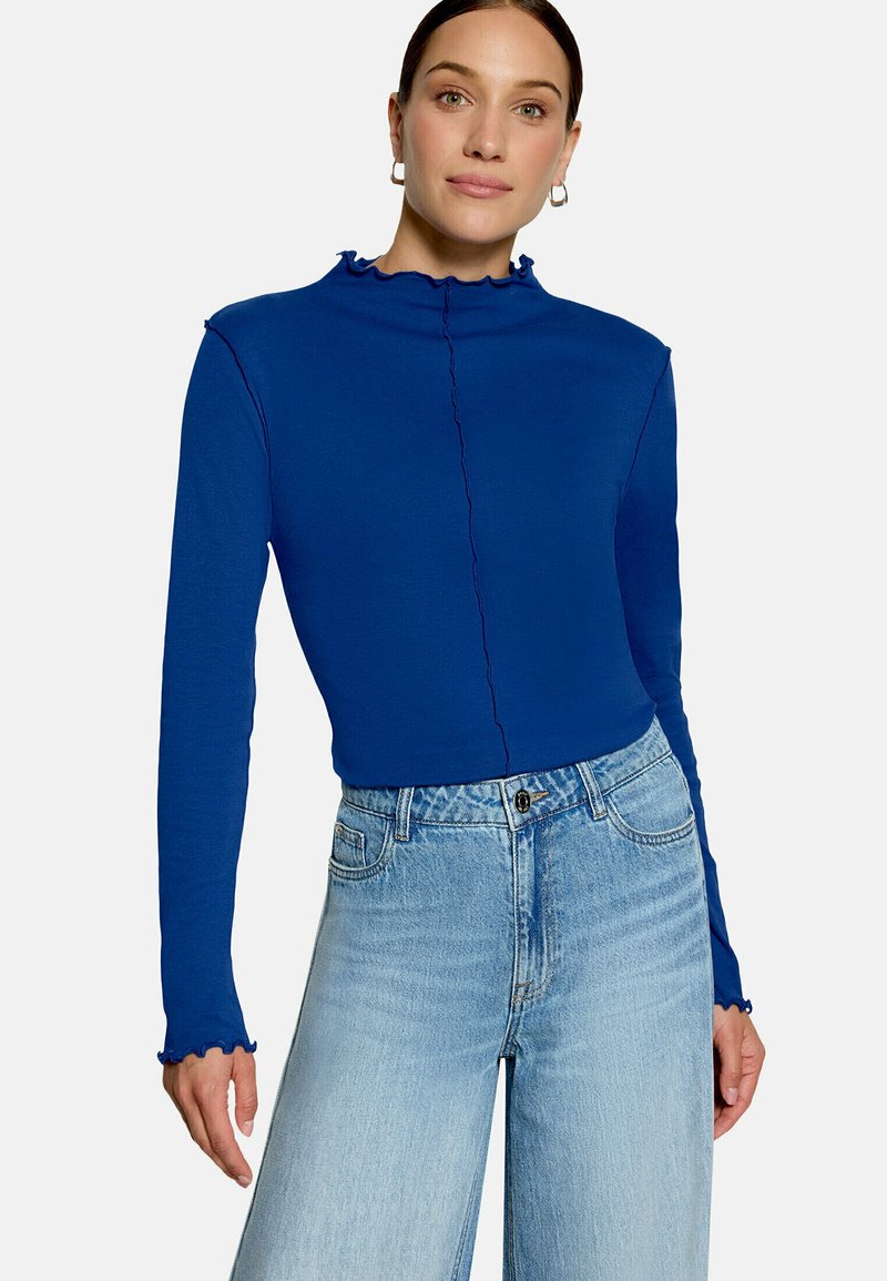 Femme portant un haut bleu ajusté à manches longues avec des bords froncés et un jean taille haute bleu clair sur un fond uni.