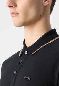 Maiou polo negru cu un guler tricotat contrastant de culoare maro, trei nasturi, țesătură texturată și un logo embosat pe piept.