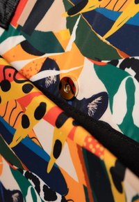 Kleurige stof met abstracte patronen in oranje, groen, geel en blauw. Heeft een zwarte rand en een gouden drukknoopdetail.