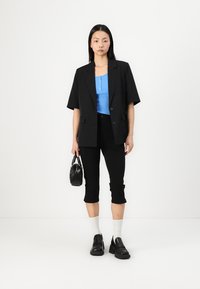 Černý oversized blazer, modrý žebrovaný top, zkrácené černé kalhoty, bílé ponožky, robustní černé boty, malá černá kabelka. Neutrální pozadí.