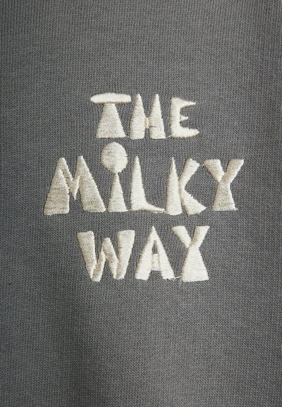 Κέντημα με τις λέξεις "THE MILKY WAY" σε λευκά στυλιζαρισμένα γράμματα μπλοκ πάνω σε γκρι πλεκτό ύφασμα.