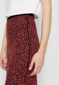 Gros plan d'une personne portant une jupe imprimé léopard rouge et noir avec un haut blanc, montrant la couture latérale et la main posée près de la jupe.