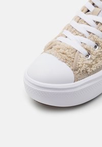 Beige sneaker med mjuk textur, vit gummitåhätta, runda skosnören och mönstrad sula. Detaljer inkluderar silverfärgade öljetter och kontrasterande kantband.