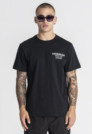 Uomo con capelli corti e tatuaggi su entrambe le braccia indossa una maglietta nera Kavanagh Winners Club e grandi occhiali da sole scuri, in piedi davanti a uno sfondo semplice.