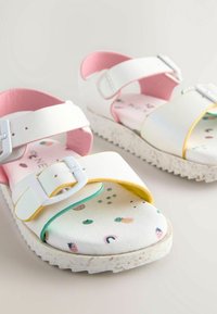 Witte sandalen met roze en gele accenten, verstelbare banden, een gestructureerde zool en een kleurrijk fruit- en regenboogpatroon op de binnenzolen.
