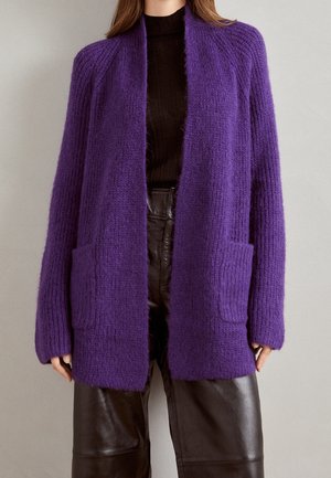 Femme portant un long cardigan violet pelucheux sur un col roulé noir côtelé et un pantalon en cuir marron foncé, debout contre un mur uni.