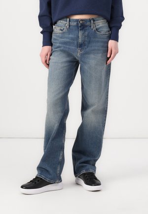 Person wearing faded blue wide-leg jeans, navy cropped sweatshirt, and black low-top sneakers with white soles standing against white background.