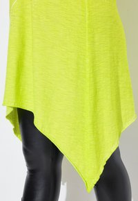 Túnica asimétrica de color verde lima con una textura suave y dobladillo deshilachado, combinada con leggings de imitación cuero negros. Acentuada con pequeños rhinestones.
