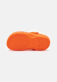 Ljust orange gummiskosula med texturerade spår och en rundad tådesign. Innehåller en cirkulär tryckknappdetalj nära remmen.