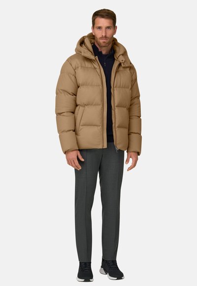 Homme portant une doudoune à capuche beige, un pantalon foncé, des baskets noires et un pull foncé sous la veste ouverte, debout devant un fond neutre.
