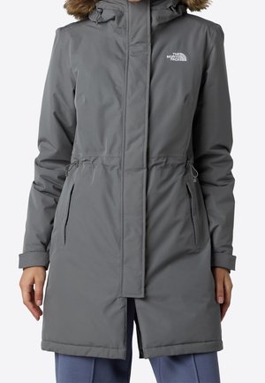 Szary zimowy płaszcz do kolan z futrzanym obszyciem kaptura, kieszeniami na zamek i logo The North Face na piersi, noszony na niebieskich spodniach.
