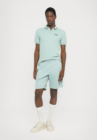 Polo verde claro com detalhe de colarinho em contraste, combinado com calções a condizer. Apresenta um discreto logótipo EA7. Estilizado com ténis bege e meias brancas.