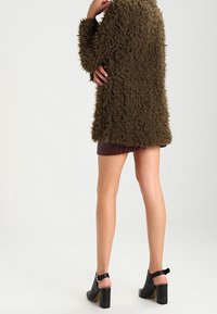 Sofiany oliwkowy płaszcz z długimi rękawami i luźnym krojem, zestawiony z bordową spódnicą i czarnymi sandałami na słupku.