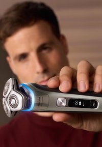 Philips scheerapparaat met een zilver- en zwarte kunststof behuizing, drie ronde scheerhoofden van roestvrij staal, een blauwe LED-ring en een digitaal display dat de batterijlevensduur aangeeft.