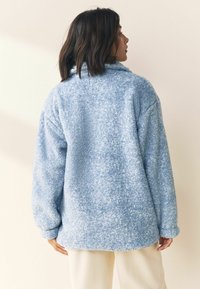 Lyseblå fleecejakke med en blød, struktureret overflade, hængende krave og løs pasform, der har lange ærmer og en rundet kant.