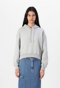 Sudadera corta de color gris claro con un bolsillo frontal y cordón. Presenta mangas largas y sueltas. Combinada con una falda de mezclilla. Fondo liso.