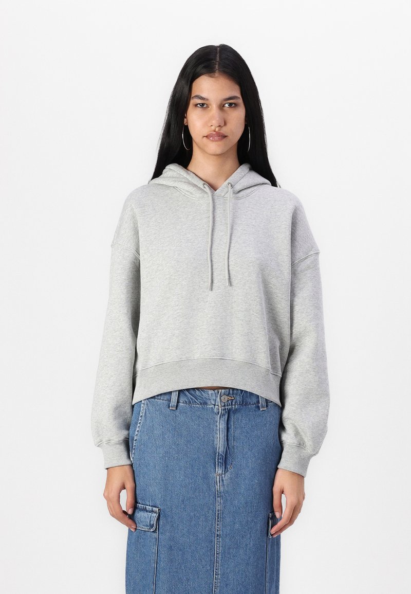 Sudadera corta de color gris claro con un bolsillo frontal y cordón. Presenta mangas largas y sueltas. Combinada con una falda de mezclilla. Fondo liso.