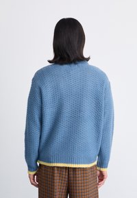 Πλεκτή μπλε ζακέτα με υφασμάτινο σχέδιο, κίτρινη κάτω βάση και μανίκια. Χαλαρή εφαρμογή με στρογγυλή λαιμόκοψη και ελαφρώς χαμηλά ώμους.