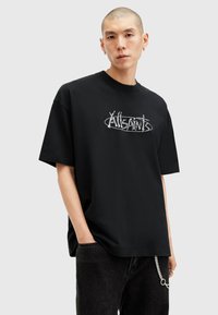 Sort kortærmet T-shirt med et hvidt grafisk logo på fronten. Båret med sorte bukser, der viser en sølvkæde som tilbehør i taljen.