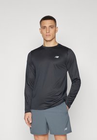 Svart långärmad träningströja i slät tyg med en subtil textur, med en liten grå logo på bröstet. Bärs med grå shorts.