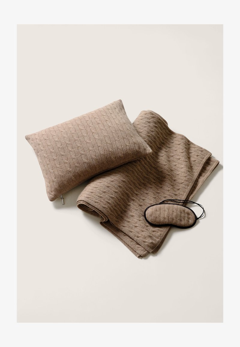 Beige stickad filt med kabelmönster, matchande kudde och en ögonmask, alla med mjuka texturer och subtila tonala variationer.