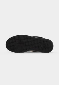 Semelle en caoutchouc noir d’une chaussure avec des motifs en grille et circulaires et un petit logo circulaire près du talon sur fond blanc.