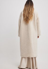 Cremefarvet lang frakke lavet af struktureret stof, med lige snit og overdimensionerede ærmer, båret over en beige nederdel.