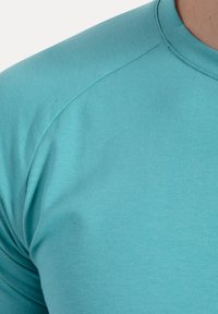 Camicia aderente turchese realizzata in tessuto morbido, con scollatura rotonda e maniche a raglan con una consistenza delicata.