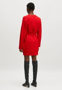 Rotes Strickkleid mit langen Ärmeln, einer taillierten Taille mit Gürtel und einem gerippten Saum. Kombiniert mit hohen schwarzen Lederstiefeln.