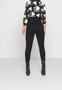 Svarta slim-fit byxor tillsammans med en mönstrad blus i svart och vitt, med långa ärmar och en subtil design, sedd bakifrån.